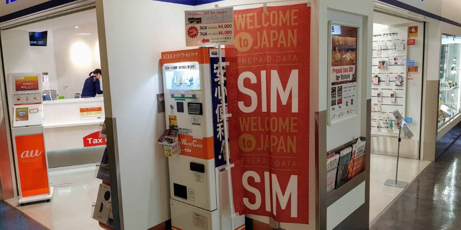 A brightly lit kiosk in a Japanese airport or shopping center with a prominent red and white banner that reads "WELCOME to JAPAN PREPAID DATA SIM." A large vending machine for prepaid SIM cards and international calling cards stands next to the banner, with prices displayed for different data plans (e.g., 3GB for ¥4,000 and 7GB for ¥6,000). A service counter with a staff member is visible in the background.