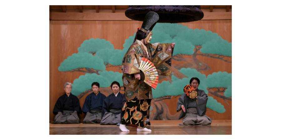A Noh performer in an elaborate brocade costume and mask stands center stage holding a folding fan. Behind them, musicians in traditional hakama sit on a wooden stage in front of a painted backdrop of a pine tree.