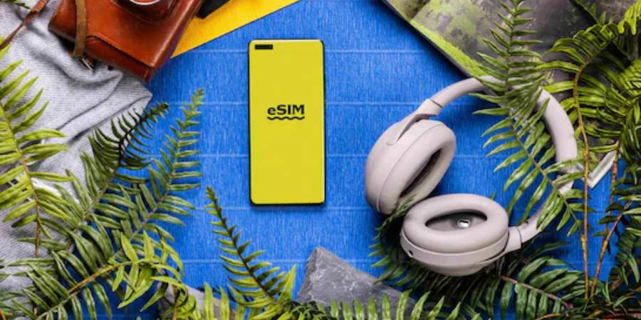 A top-down lifestyle photograph of a yellow smartphone with the text "eSIM" on its back, centered on a blue textured surface. It is surrounded by traveller's gear, including grey over-ear headphones, a brown leather camera case, a grey sweater, and green fern fronds.
