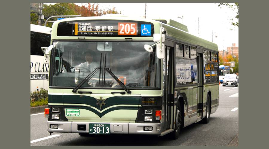 Front view of a Kyoto City Bus (Route 205) travelling on a city street, displaying the destination 'Kyoto Sta. via Nishioji Shijo' on its digital marquee.