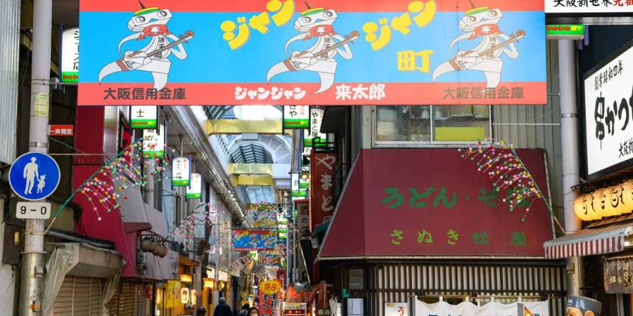 Close-up view of the entrance to Jan Jan Yokocho Alley, featuring a large banner with a caricature mascot playing a shamisen, with Japanese signs for food and shops below.