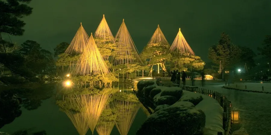 Kenrokuen Garden in Kanazawa illuminated at night in winter, featuring traditional yukitsuri rope supports on pine trees, glowing garden lights, and reflections in a pond.