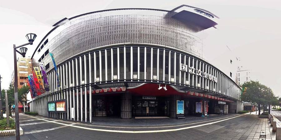 Wide panoramic view of the Osaka National Bunraku Theater, featuring its distinctive facade of vertical black and white pillars and wide, curved structure.