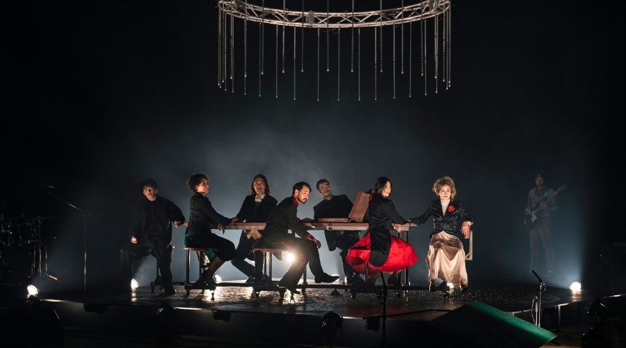 A wide stage shot of seven actors seated at a long table under a circular light fixture with hanging crystal-like strands. The stage is dark and atmospheric with low-level fog, and the cast is dressed in formal or semi-formal attire, including one woman in a striking red skirt.