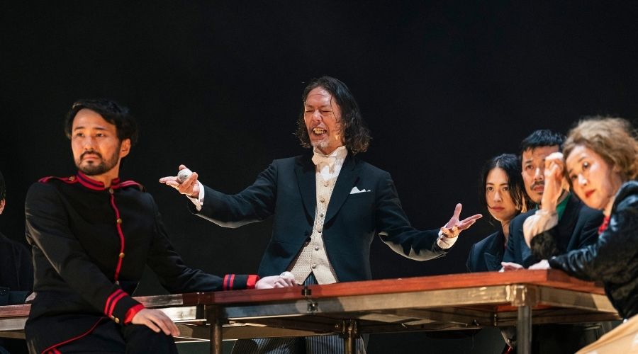 A collage of three vertical images showing theatrical scenes. Actors are shown in dramatic poses around a wooden table under hanging light fixtures, with one actor appearing to reach toward the ceiling.