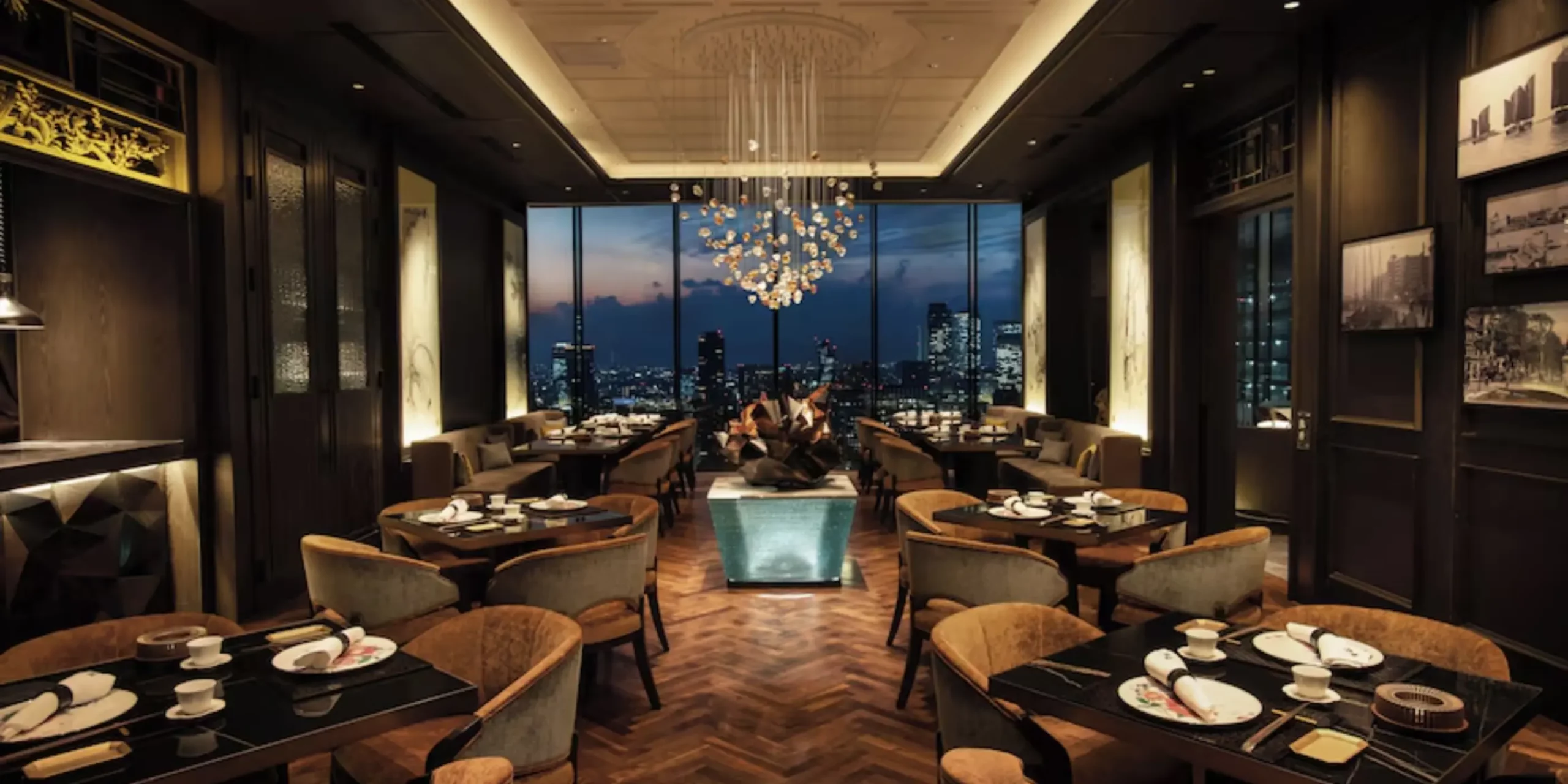 Elegant restaurant interior at The Royal Park Hotel Iconic Nagoya, featuring neatly set tables, a central chandelier, and floor-to-ceiling windows overlooking the city skyline at dusk