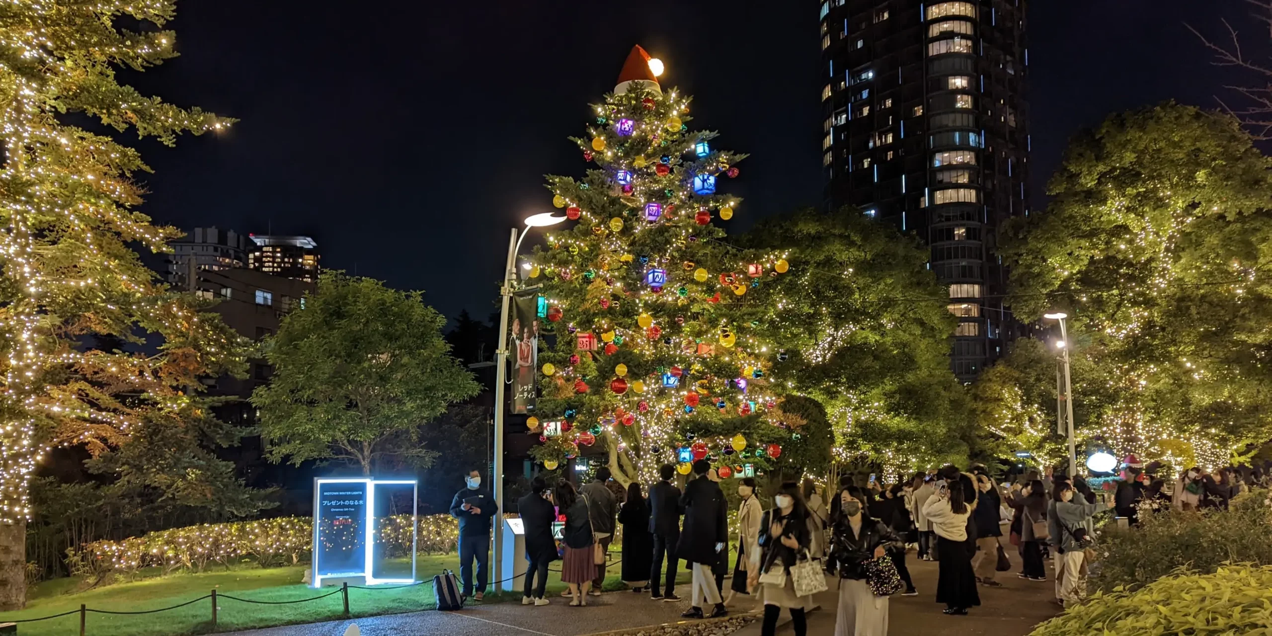 Tokyo Midtown Christmas scene with a decorated tree glowing in multicoloured lights, surrounded by illuminated trees and evening crowds enjoying the festive display