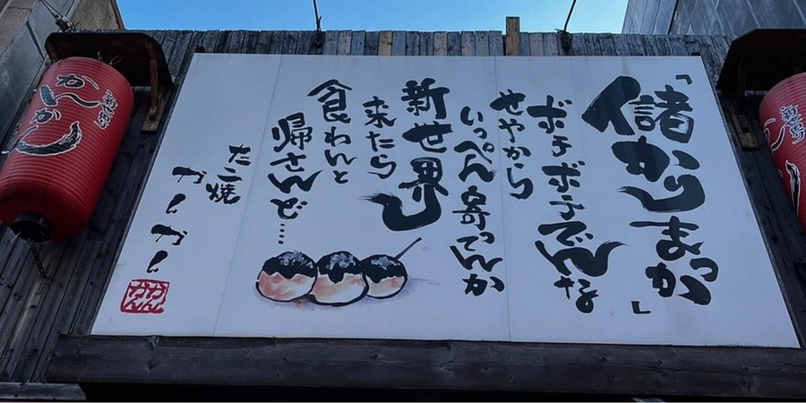 Large storefront sign in Shinsekai with bold Japanese calligraphy and an illustration of takoyaki, flanked by red lanterns on either side