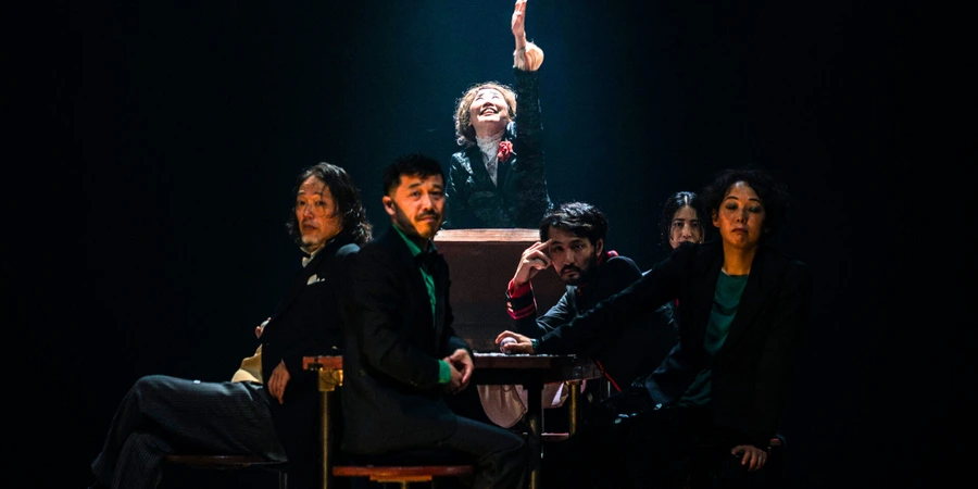 Performers of The Gambler sit and stand around a small table under a single spotlight, dressed in dark suits; one figure rises at the centre with an arm raised, creating a tense, theatrical tableau against a black stage backdrop.