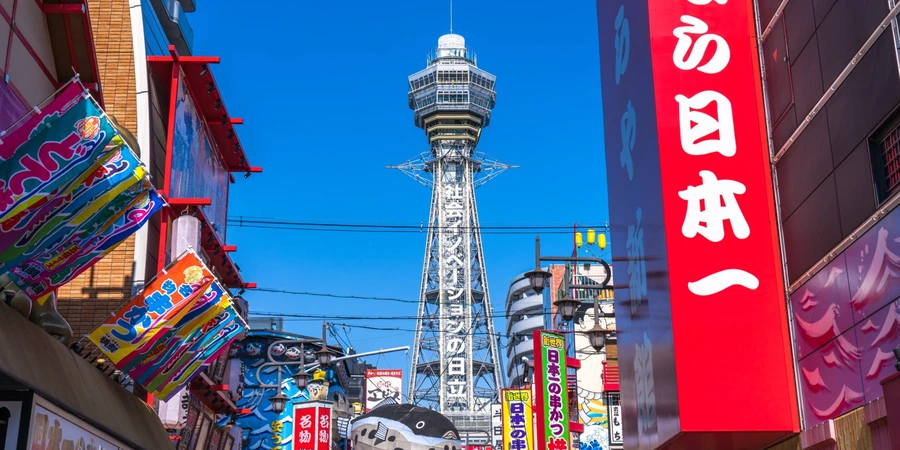 Tsutenkaku Tower rising above Shinsekai in Osaka during the day, framed by colourful shop signs and busy street storefronts
