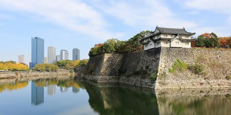 Osaka Castle moat reflecting stone ramparts and a corner turret, with autumn trees and the city skyline in the background