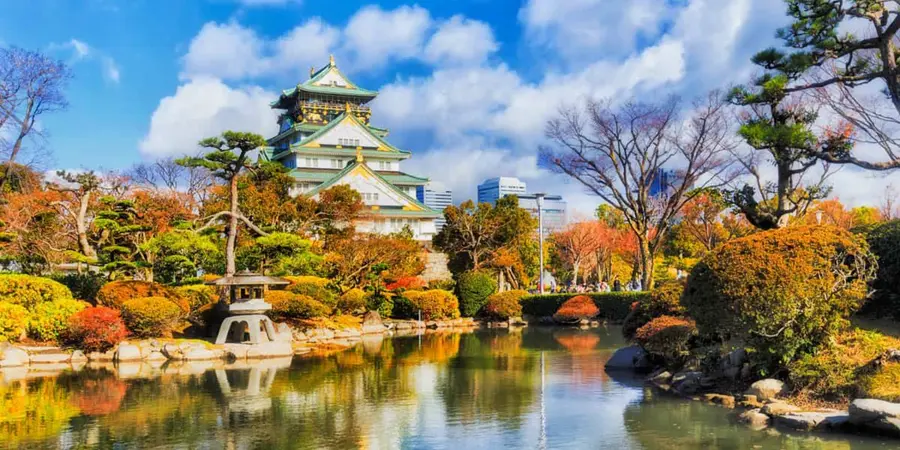 Osaka Castle overlooking a tranquil garden pond, framed by autumn foliage, sculpted pines, and stone lanterns under a bright blue sky
