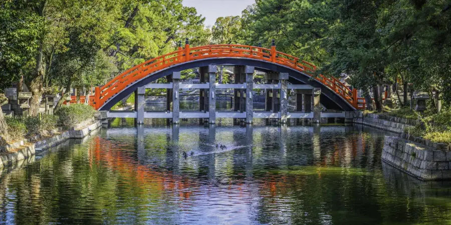 Arched vermilion bridge at Sumiyoshi Taisha spanning a reflective canal, with stone embankments and dense greenery framing its vivid reflection in calm water.
