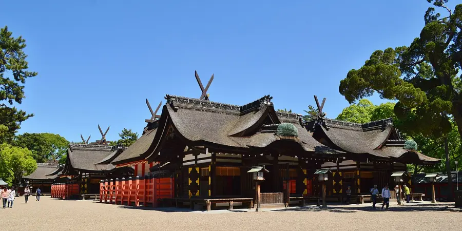 Low-angle view of Sumiyoshi Taisha’s main shrine (honden), showing sweeping thatched roofs with crossed chigi finials, red railings, and visitors walking across the gravel courtyard under a clear blue sky.