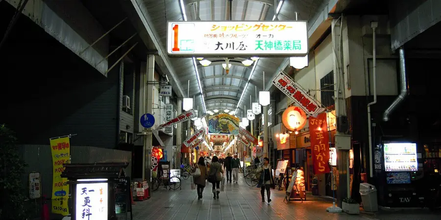 Tenjinbashisuji Shopping Street at night, lit by shop signs and lanterns under the covered arcade, with pedestrians walking through the lively Osaka corridor