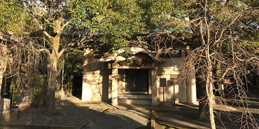 Shaded entrance to Sumiyoshi Taisha’s Treasure Museum, a small plaster-walled building framed by trees, with dappled sunlight across the gravel path.
