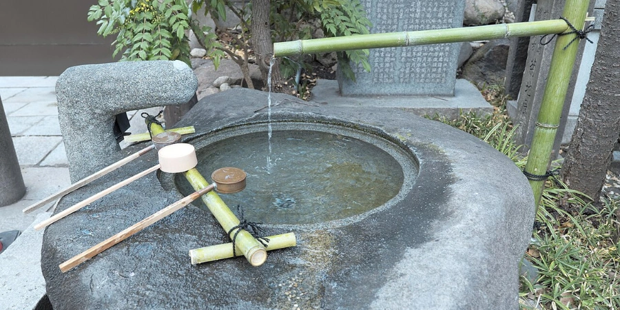 Stone chozuya basin at Namba Yasaka Shrine Osaka filled with clear spring water and a wooden dipper (hishaku) for Shinto purification ritual, surrounded by bamboo ladle rests and pebble gravel in the shrine's outer courtyard.