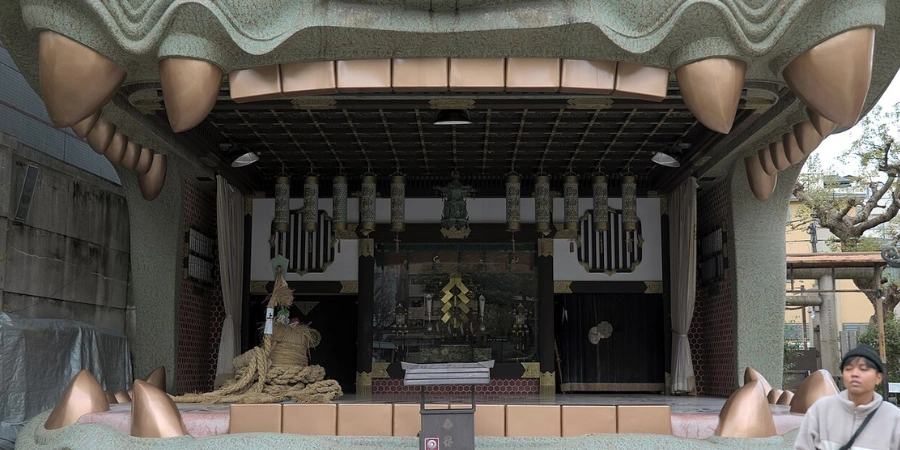 Dramatic close-up of the ceremonial lion head stage (Shishi-den) at Namba Yasaka Shrine Osaka, showcasing sharp golden teeth, and steel-concrete structure rebuilt in 1974, designed to "swallow evil spirits" with a door inside the 10-meter-deep open mouth.