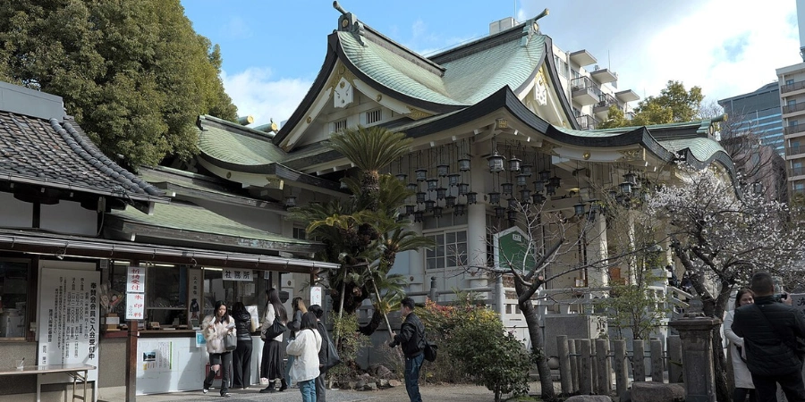 Traditional wooden main prayer hall (haiden) at Namba Yasaka Shrine Osaka, featuring a gabled roof, offering box, and other smallers buildings flanked by stone lanterns in the compact courtyard near the lion head structure.