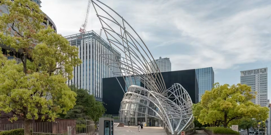 Striking inverted pink tetrahedral exterior of the National Museum of Art, Osaka, partially embedded in Nakanoshima ground with curved glass ramps leading underground, contrasting the surrounding park greenery and river.