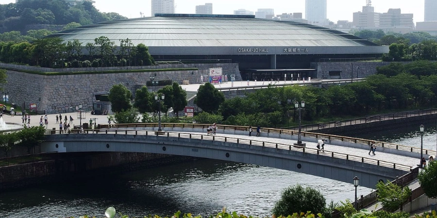 Panoramic view of sail-shaped Osaka Jo-Hall exterior amid expansive Osaka Castle Park greenery, steps from the Osaka Museum of History, showcasing event venue architecture and seasonal cherry blossoms.