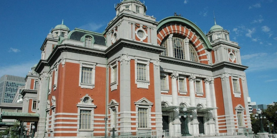 Majestic red-brick Edwardian Baroque facade of Osaka Central Public Hall (1918), with ornate clock tower, arched windows, and copper dome rising above Nakanoshima plaza amid cherry trees and urban park setting.