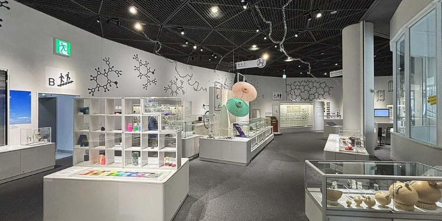 Dynamic 3F "Exploration of Matter" at Osaka Science Museum, showing visitors handling crystals, testing metal conductivity, mixing colors in chemistry stations, and watching live demos on a science stage amid material sample displays.