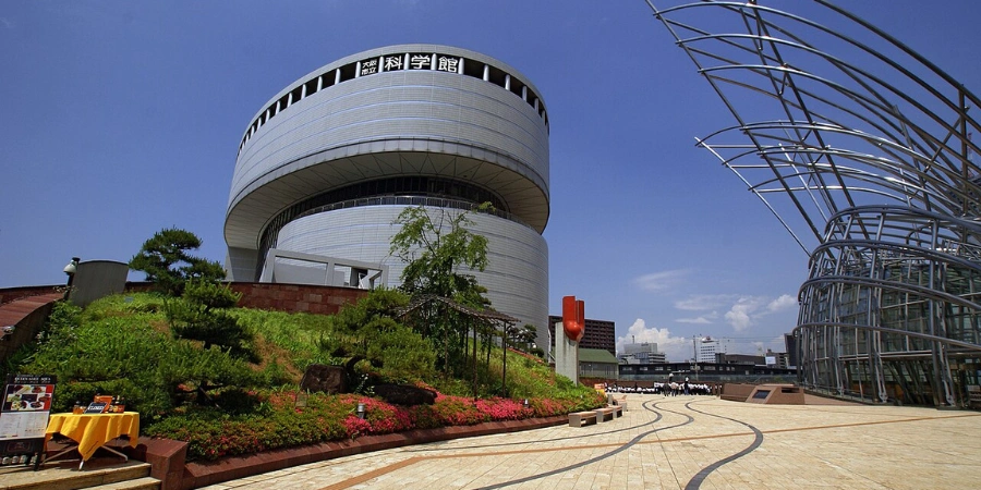 Iconic exterior entrance of Osaka Science Museum on Nakanoshima Island, showcasing the distinctive white cube structure with a gleaming silver dome against the riverside skyline and Yodogawa River bridges.