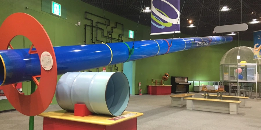 Lively sound experimentation zone on 2F at Osaka Science Museum, with curved parabolic reflectors, giant echo tubes for whispering, vibrating Kundt's tubes with dancing beads, playable global instruments like cajons and pipes, and wave visualizers.