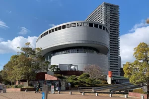 The exterior building of Osaka Science Museum. It consists of a round shape with a tall vertical building beside it.