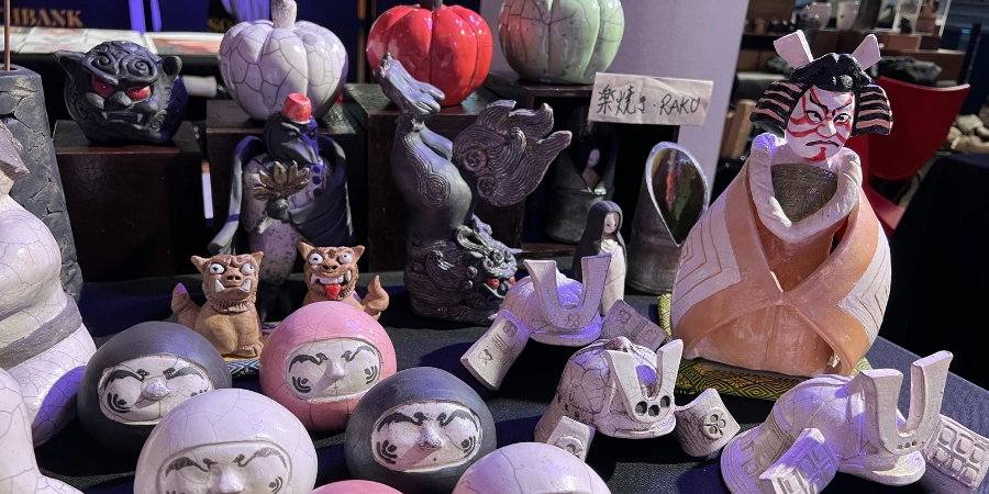 Display of handmade Japanese ceramic Daruma dolls and figurines at a market stall, featuring traditional designs and various shapes and expressions.