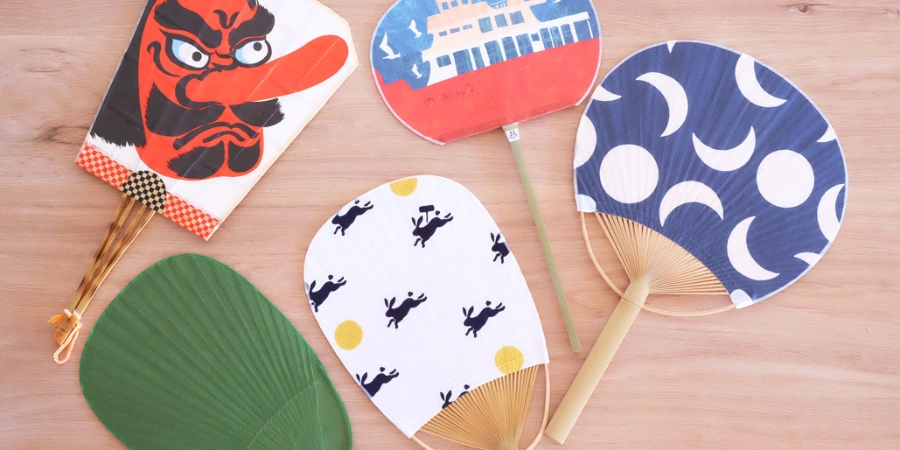 Flat lay of traditional Japanese uchiwa hand fans with colorful patterns and designs, including a Daruma motif and seasonal illustrations on a wooden surface.