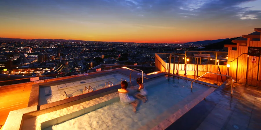 Rooftop onsen hot spring bath at sunset overlooking Osaka city skyline with people relaxing in warm water