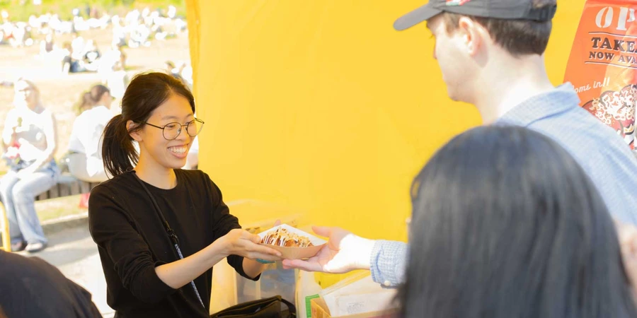 Vendor handing a freshly prepared order of takoyaki to a customer at a Japanese street food stall during an outdoor festival.