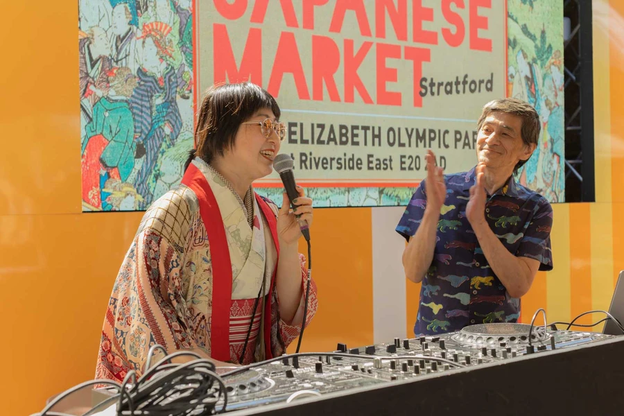 Woman in traditional kimono speaking into a microphone during a live performance at Yokimono Japanese Market in Stratford, London, with a person applauding beside a mixing deck at Queen Elizabeth Olympic Park.