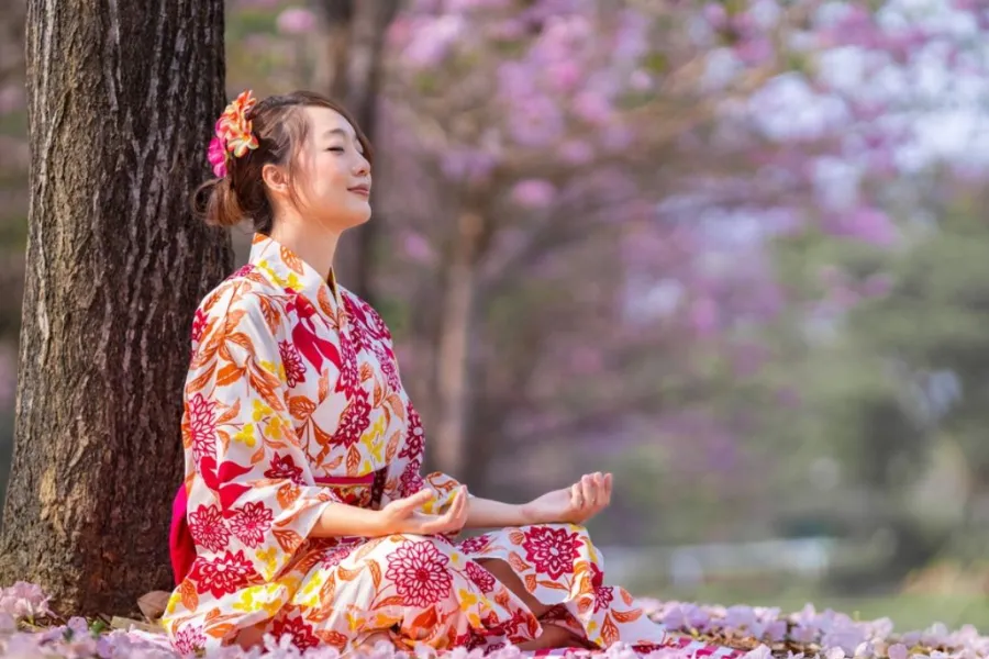 Woman meditating in a floral yukata beneath blooming cherry blossom trees in Japan, enjoying a peaceful spring hanami experience in a tranquil park.