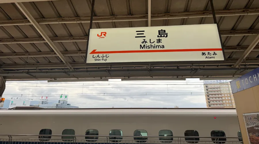 JR Mishima Station sign above the Shinkansen platform in Shizuoka Prefecture, Japan, showing directions toward Shin Fuji and Atami with a bullet train visible below.