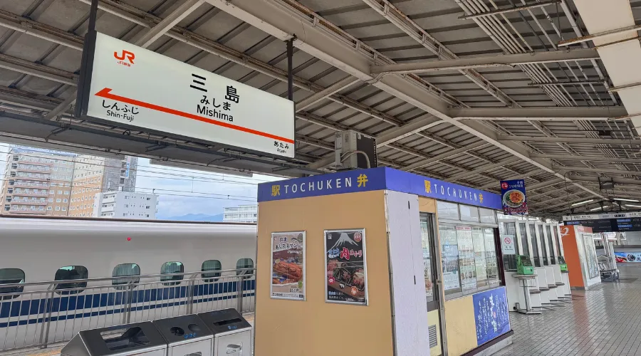 Wide view of Mishima Station Shinkansen platform in Japan with JR station signage, a Tōchūken bento kiosk, and a parked bullet train alongside the platform.
