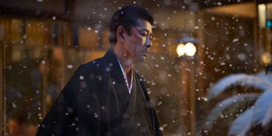 Cinematic still from Kokuho featuring a wounded yakuza character standing in falling snow outside a traditional Japanese building at night