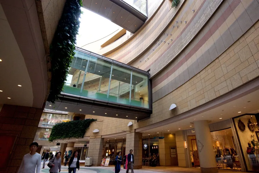 Interior view of Namba Parks Shopping Complex in Osaka, Japan, featuring curved modern architecture, open-air atrium, glass skybridge, retail storefronts, and shoppers walking through the luxury shopping mall.