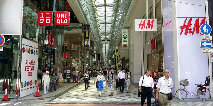 Busy Shinsaibashi-suji shopping arcade in Osaka, Japan, featuring popular fashion retailers including Uniqlo and H&M, covered pedestrian street, modern storefronts, and shoppers walking through the vibrant retail district.