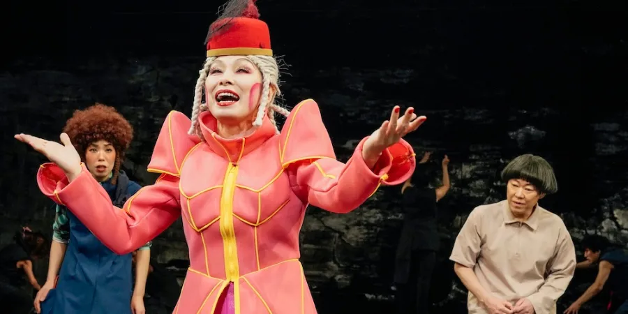 Actor in a vibrant pink costume performing on stage during the 320°F Play theatre production, surrounded by performers in a colourful contemporary theatrical scene