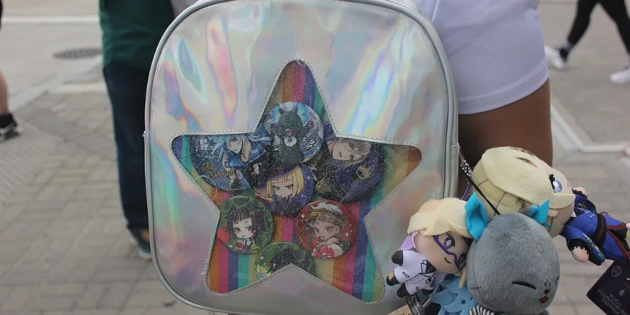 Ita bag decorated with Disney Twisted Wonderland character badges and plush keychains inside a holographic backpack at an anime event.