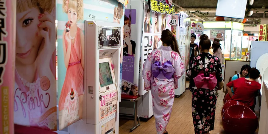 Visitors wearing colourful yukata walking through a Japanese arcade filled with purikura photo booths and kawaii gaming attractions.