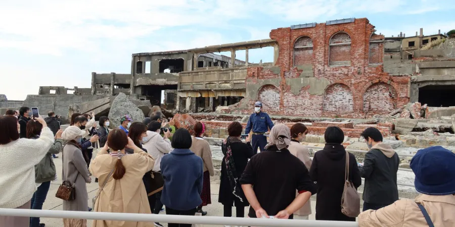 Visitors on a guided Gunkanjima Island tour viewing the ruins of abandoned industrial buildings and red brick structures.
