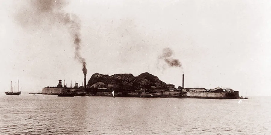 Historic black-and-white photo of Hashima Island during its coal mining era, with industrial smoke rising from the island.