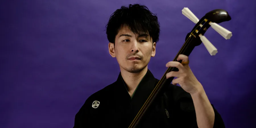 Hibiki Ichikawa in traditional black attire performing against a purple backdrop during a live cultural music performance.