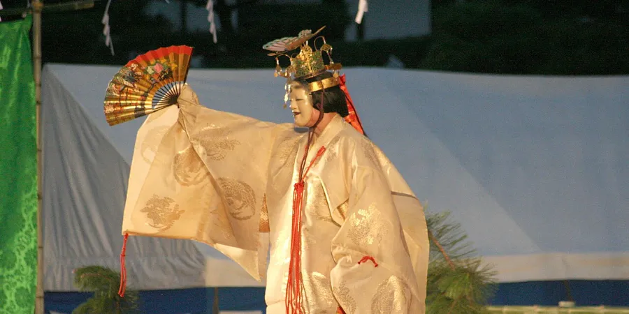 Takigi Noh performer in traditional white costume holding a folding fan during a Japanese theatre performance.