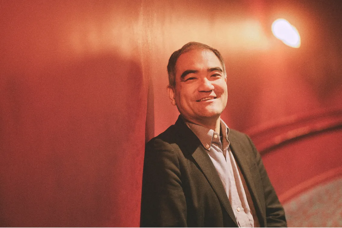 Composer Dai Fujikura smiling in a theatre, illuminated by warm red and orange lighting.