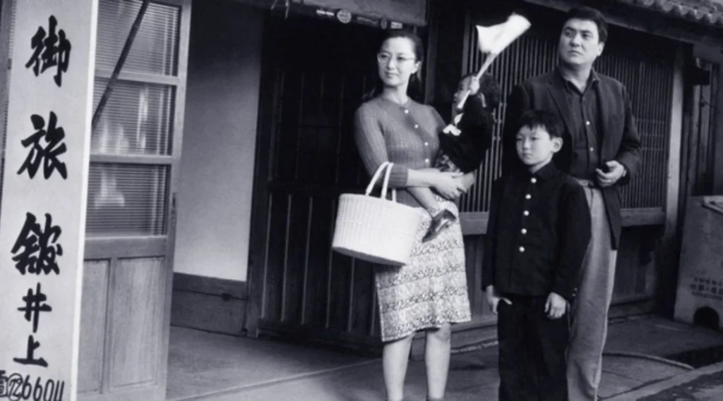 The family from Boy (1969) standing outside a Japanese inn: the stepmother holding the toddler, the Boy in his school uniform and the father beside them